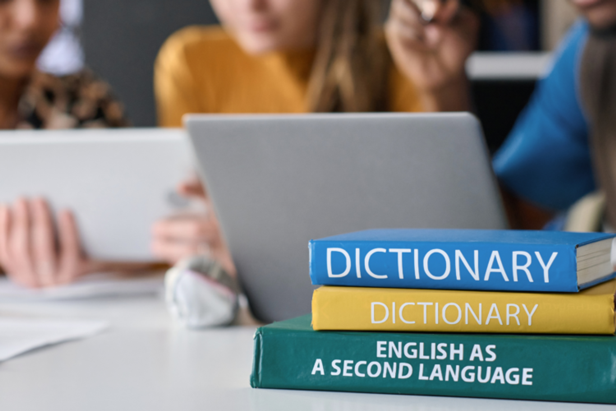 People working at a table on laptops beside a pile of dictionaries and a book titled "English as a second language"