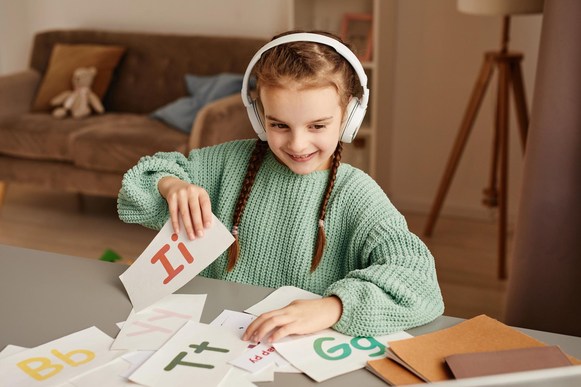 A girl with headphones looking at a screen, she is holding coloured cards, each one with a letter of the alphabet in upper and lower case..