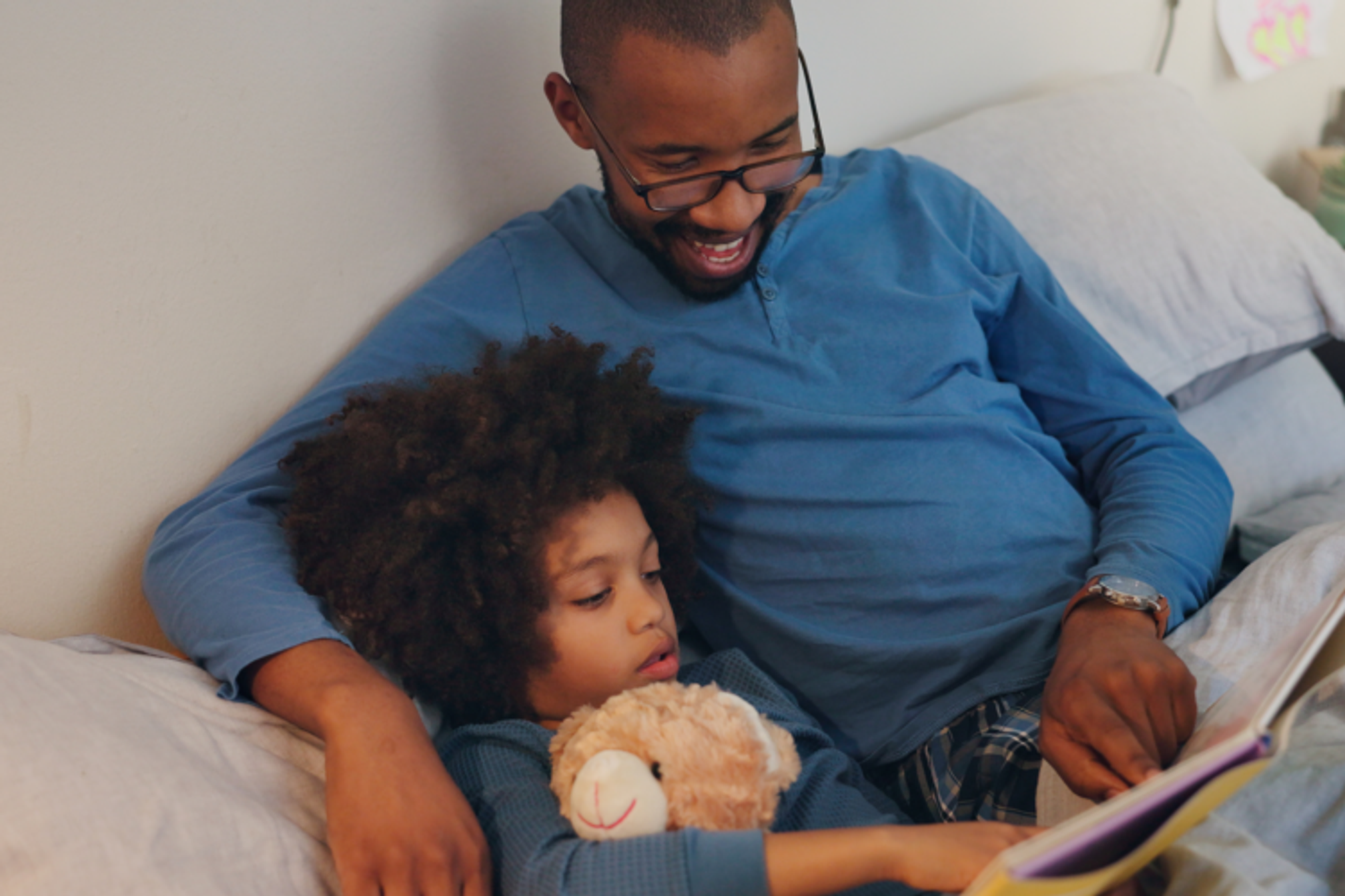 A young man reading a book with his child in bed.