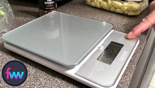 Kindal is setting setting her food scale up and pointing to the tare button. This button will make tracking your meals very easy and much less of a headache. 