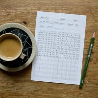 A half filled in coffee tasting card next to a cup of espresso and a pencil on a wooden surface.