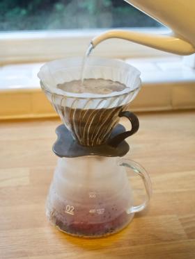 Hario's V60 with a gooseneck kettle pouring hot water into the cone full of fine coffee. Brewed coffee drips into the carafe below.