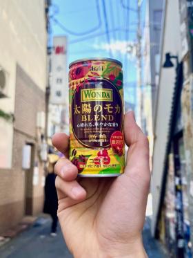 A can of Wonda Taiyō no Moka Blend held up against the background of a narrow street in Tokyo