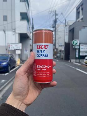 A can of UCC milk coffee held up against the backdrop of a street and parking lot