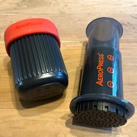 A black translucent plunger with the Aeropress logo next to a black plastic cup with a bright red rubber lid.