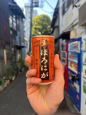 A can of Boss Coffee Horoniga held up against the backdrop of an alley way filled with vending machines