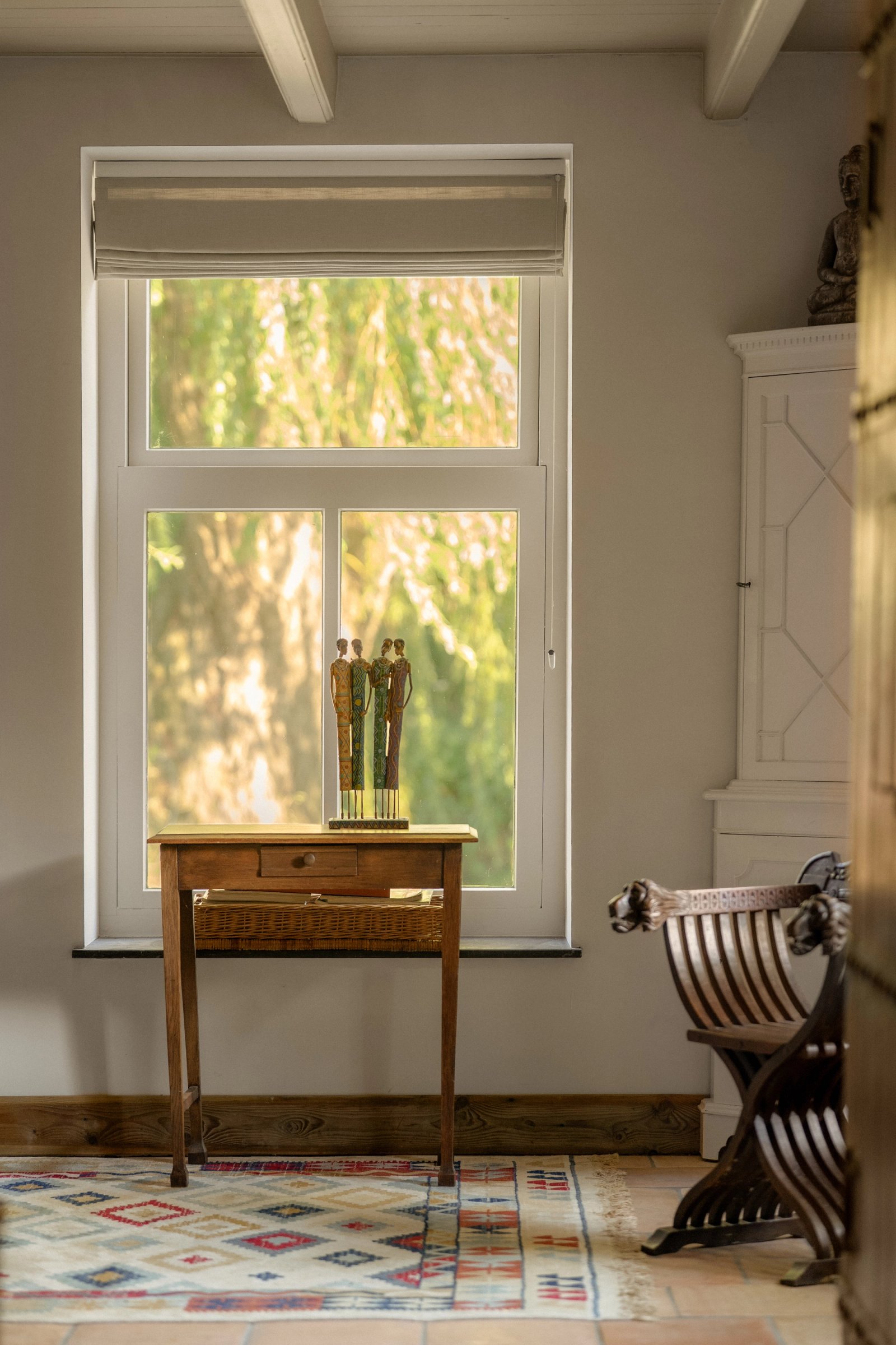 Hoog raam met uitzicht op groen, een houten bijzettafel en een klassieke stoel in zacht daglicht