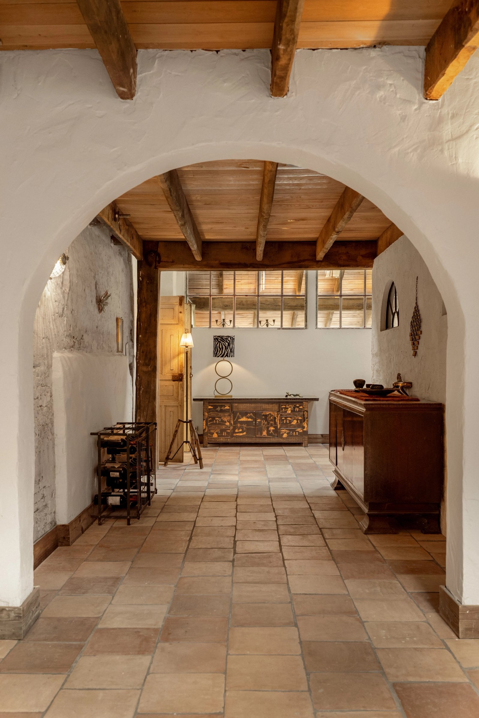 Doorkijk door een witte toogboog naar een hal met houten balkenplafond en terracotta vloer