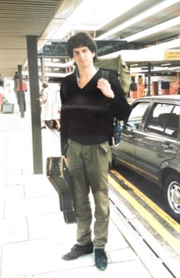 Rob as young man standing at airport with battered guitar case and rucksack