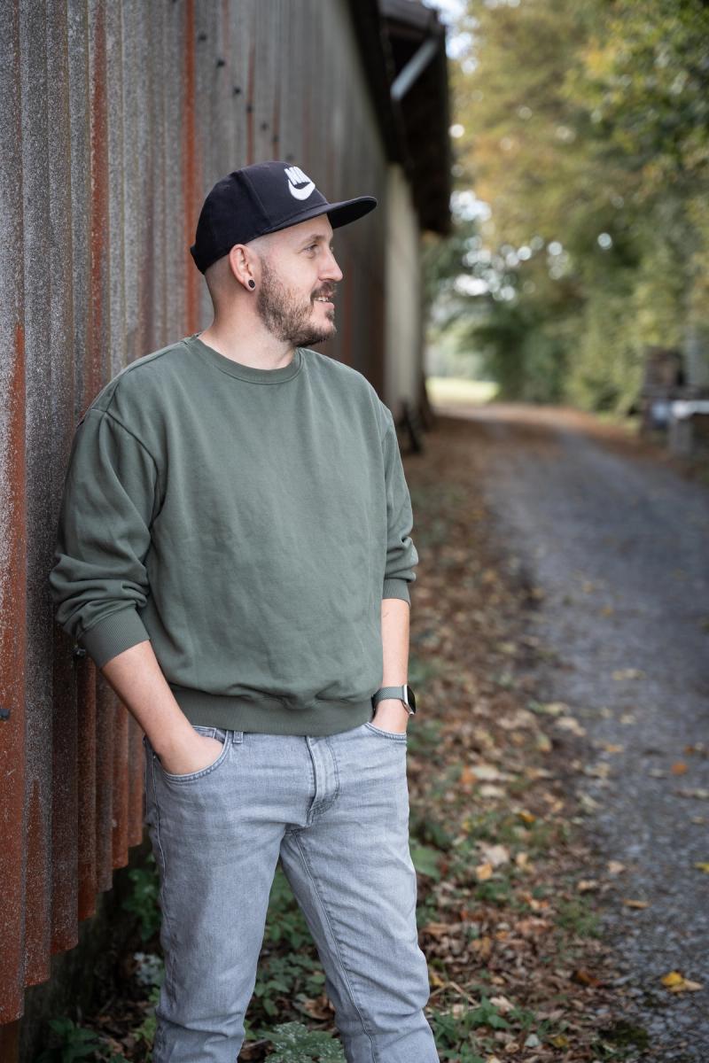 Benny Schey steht an einer verwitterten Scheunenwand und schaut zur Seite, mit Basecap und olivgrünem Pullover