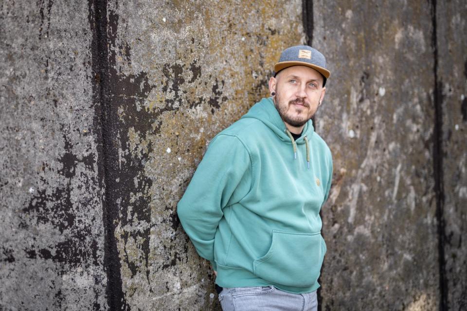 Benny Schey lehnt an einer alten Betonwand, mit Basecap und mintgrünem Hoodie