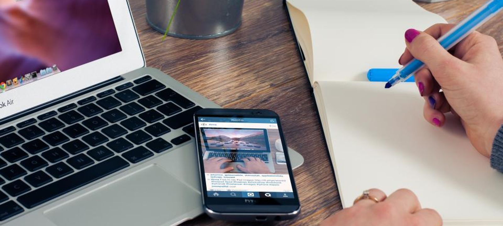 Laptop, Smartphone und Notizbuch auf einem Schreibtisch, eine Hand schreibt
