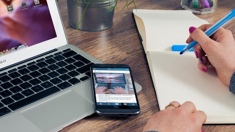 Laptop, Smartphone und Notizbuch auf einem Schreibtisch, eine Hand schreibt