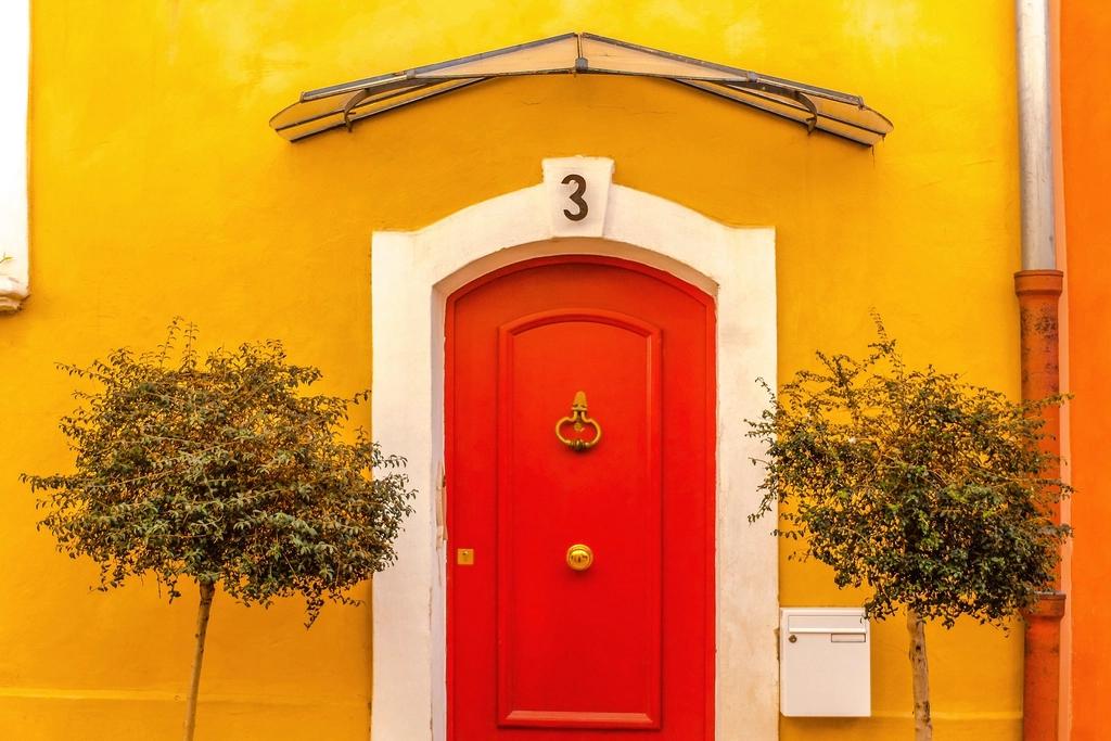 Rote Haustür in einer gelben Hauswand mit zwei Bäumchen