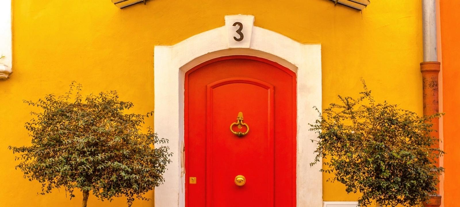 Rote Haustür in einer gelben Hauswand mit zwei Bäumchen