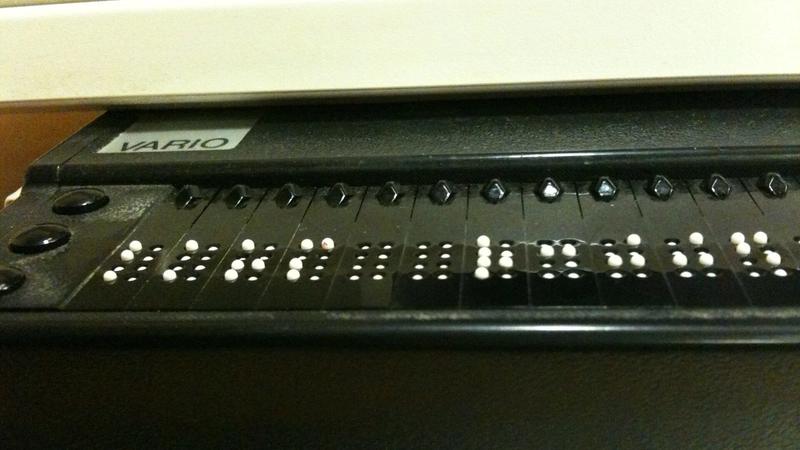 Nahaufnahme einer Braillezeile unter einer Computertastatur, die erhabenen weißen Punkte bilden Buchstaben in Blindenschrift