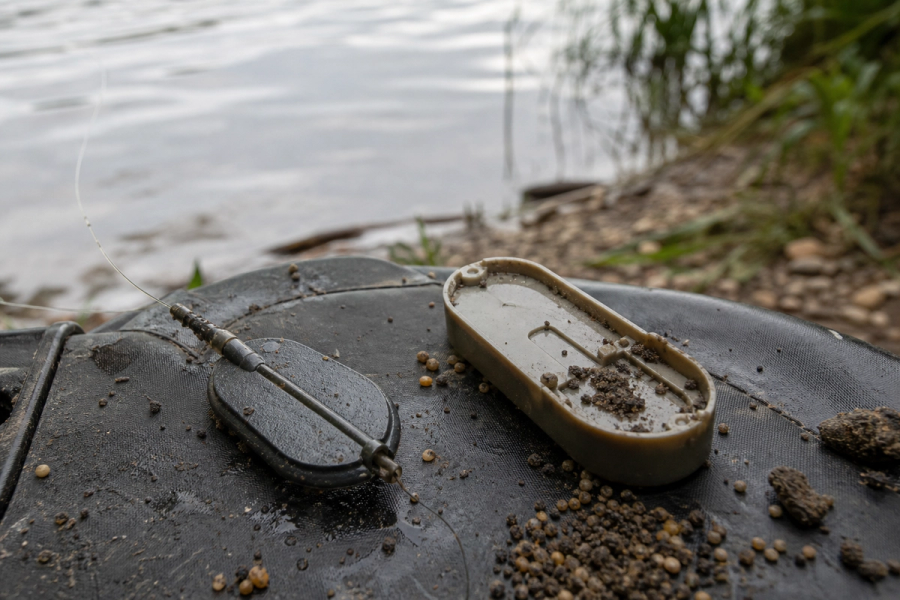 Flat inline method feeder and matching mould resting on a dark unhooking mat beside water