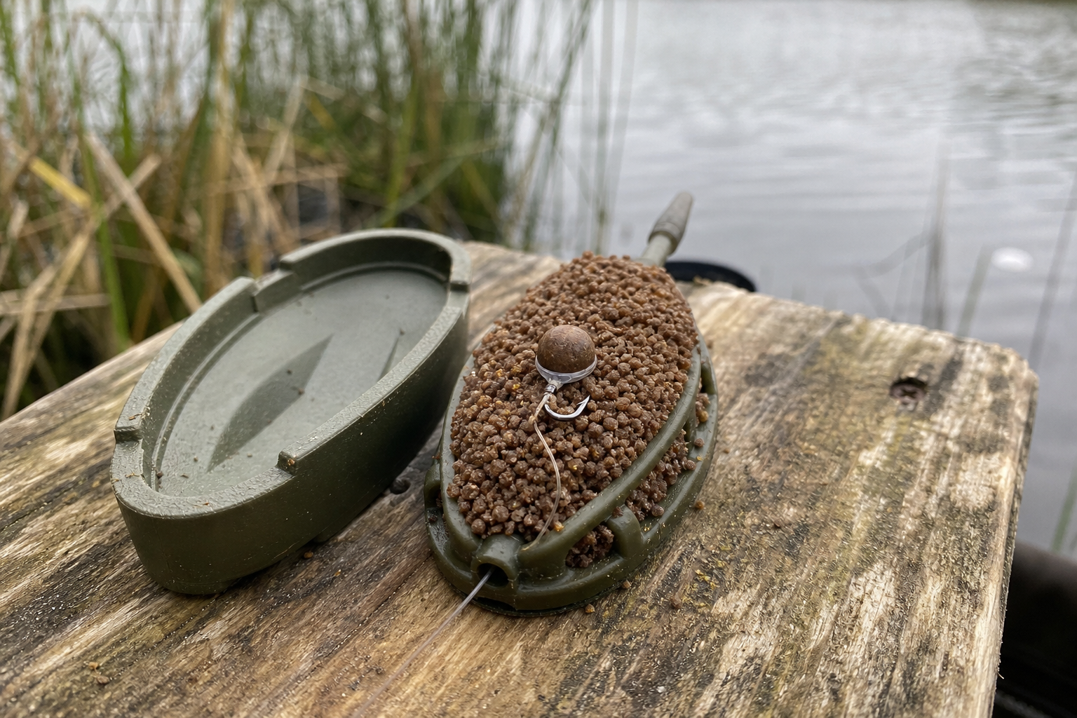 A flat-bed method feeder freshly loaded from its mould on a wooden fishing platform, with a compressed ball of micro pellets packed into the frame and a banded hard pellet hookbait pressed into the surface, the empty green mould beside it and reeds and lake water behind