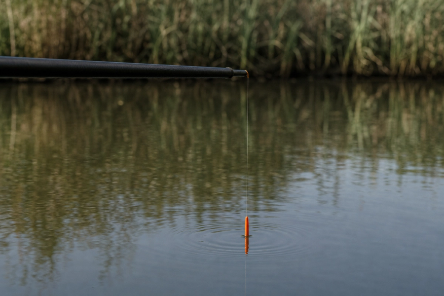 The tip of a black carbon fishing pole held out over a calm canal, with a single fine line running straight down to a small orange-tipped float and ripples on the water, reeds along the far bank