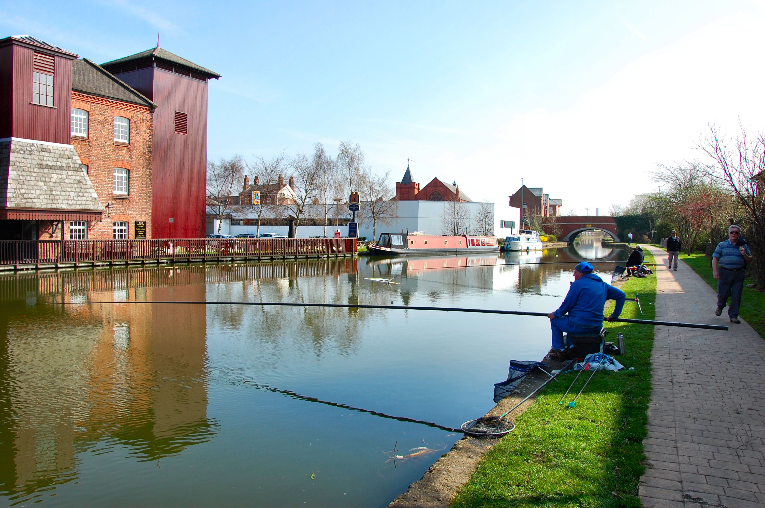 Anglers on a canal towpath holding long fishing poles out across the water, with no reels, beside a red-brick mill in Leigh