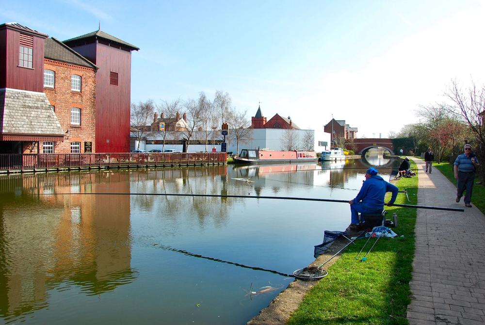 Anglers on a canal towpath holding long fishing poles out across the water, with no reels, beside a red-brick mill in Leigh