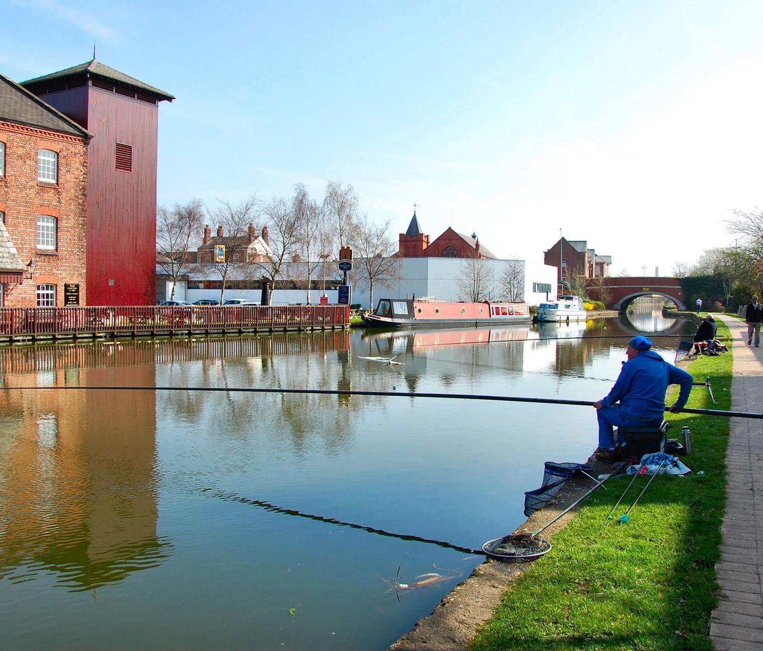 Anglers on a canal towpath holding long fishing poles out across the water, with no reels, beside a red-brick mill in Leigh