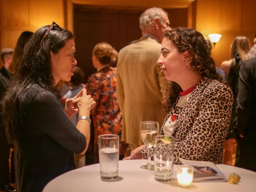 A photo of two women talking. Andrea, on the left, wears a black cardigan and glasses on top of her head, with her index fingers touching like she is counting. Kate, on the right, wears a cheetah-print cardigan and a white t-shirt with a red collar.