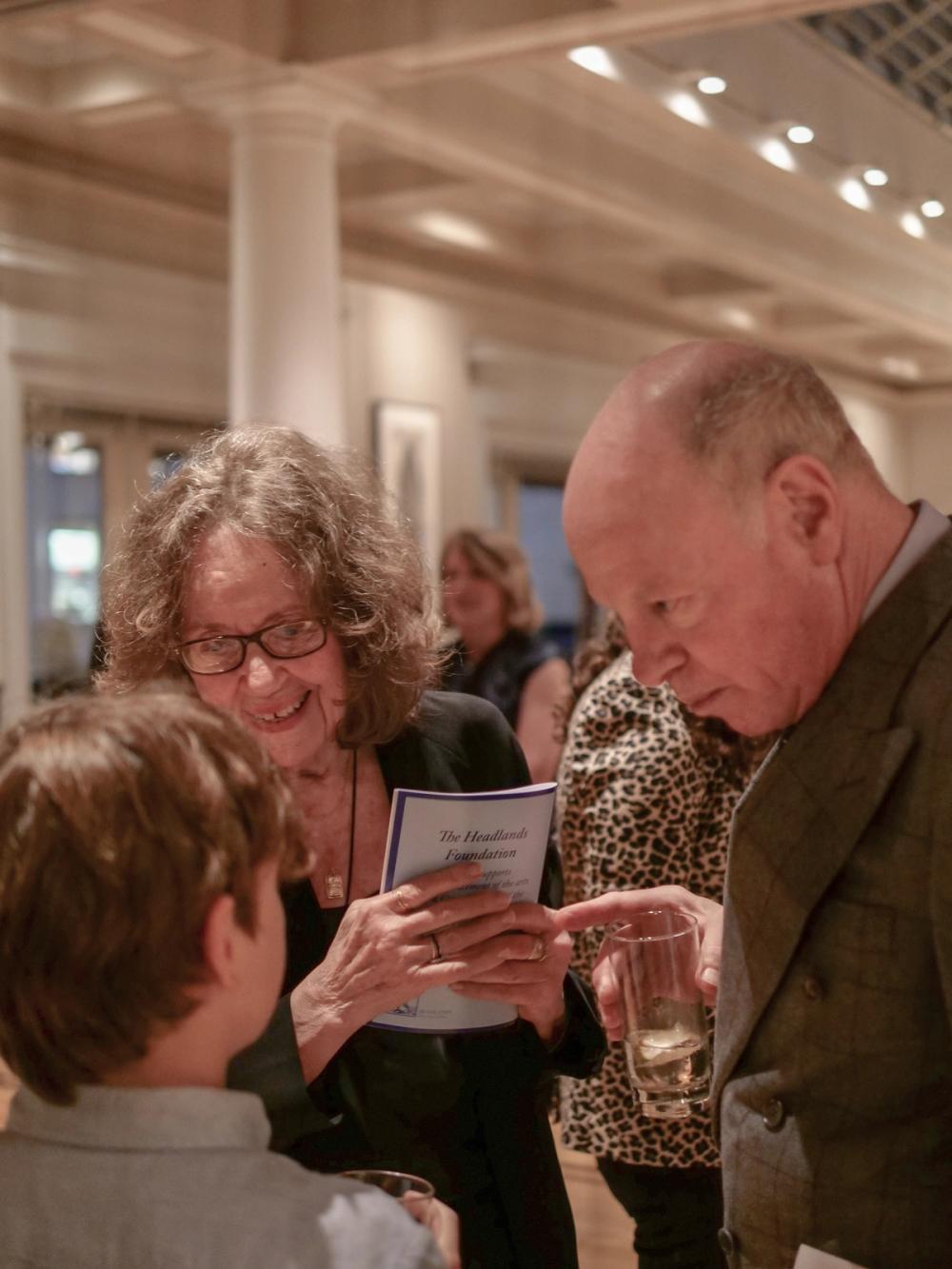 A photo of two people speaking to a young boy. Prudence, left, holds a brochure in her hand and wears dark rectangular glasses. Nick, on the right, wears a grey jacket and holds a glass in his right hand.