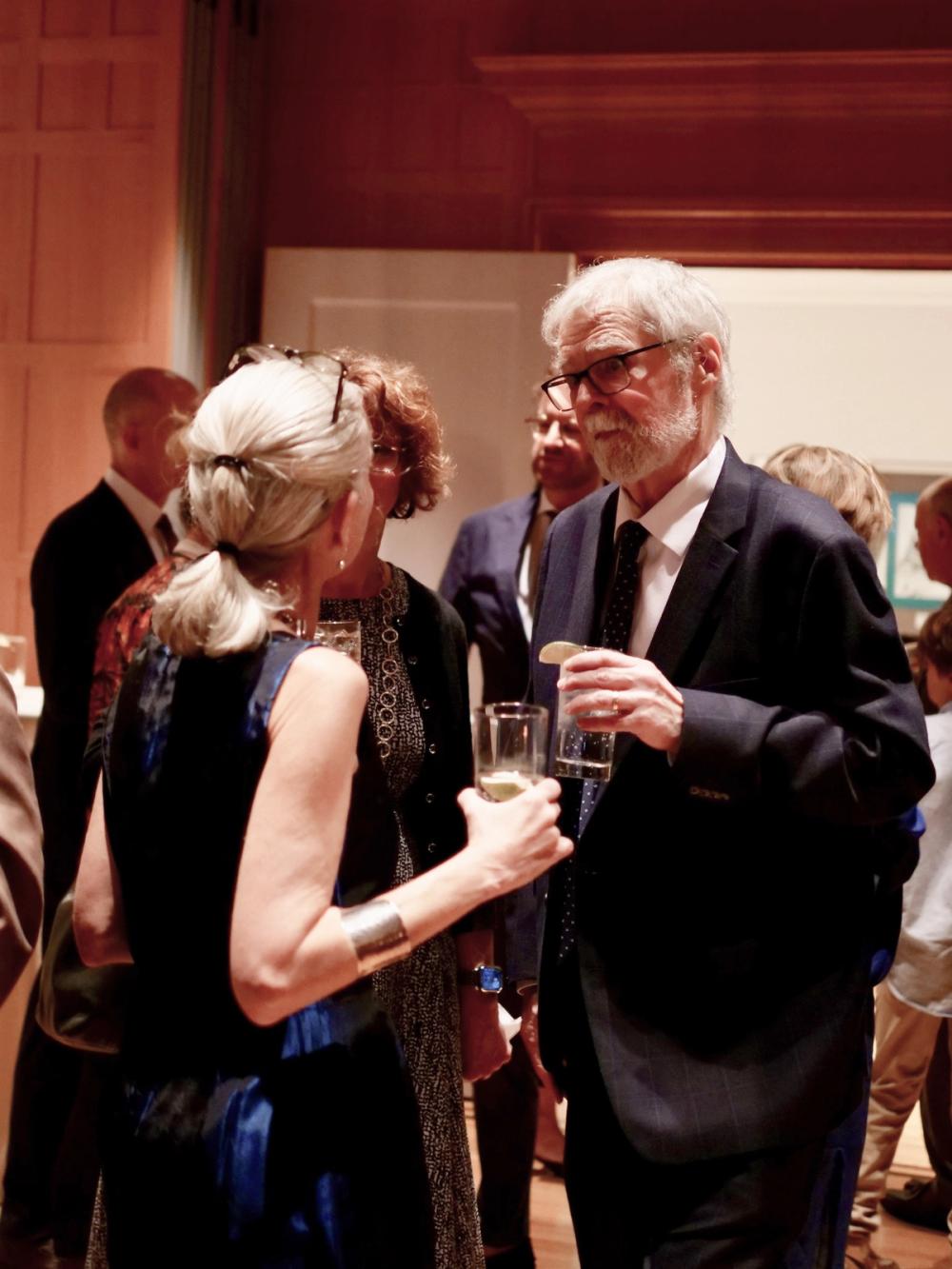 A photo of two people talking. Erika, with her back to the camera, is wearing a blue dress and holds a glass in her right hand. Joe, on the right, is wearing a dark blue suit and holds a glass in his left hand.