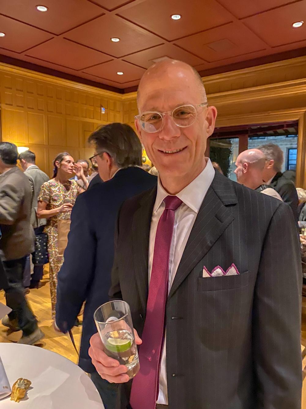 A photo of a man smiling at the camera. William wears a black striped suit and a purple tie with a matching pocket square. He holds a glass in his right hand.