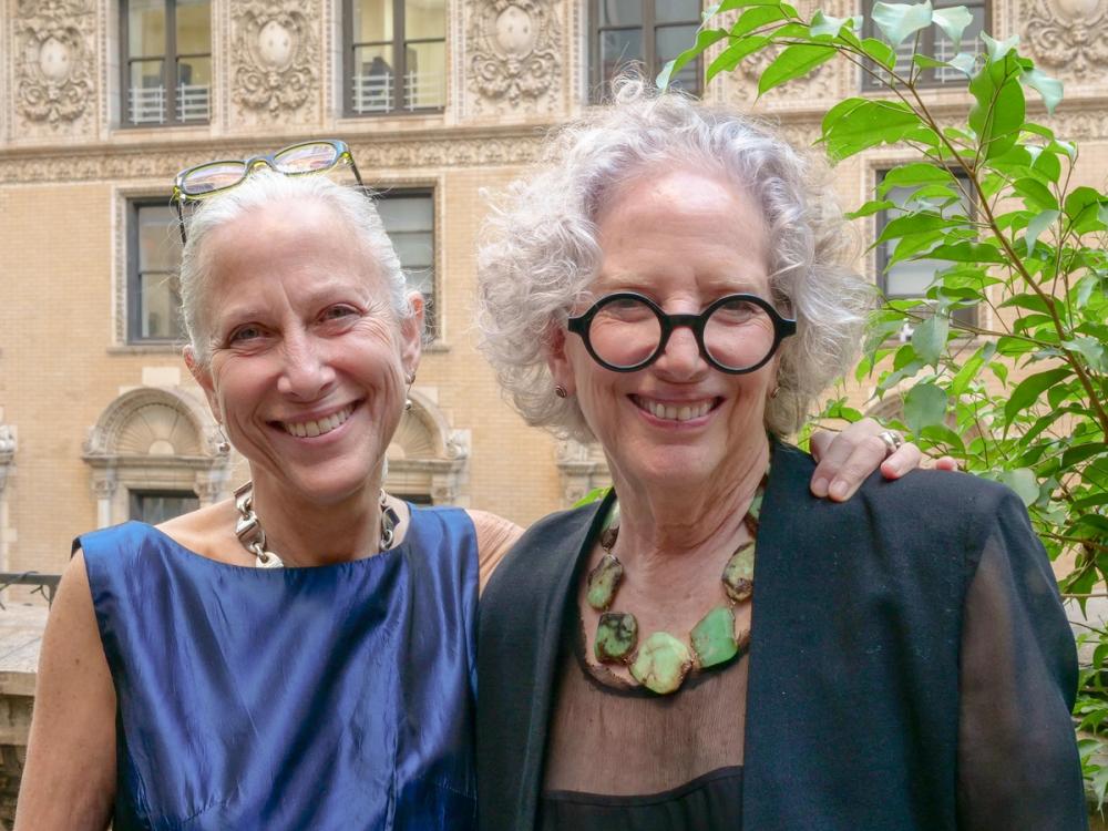 A photo of two women smiling at the camera. Erika, left, wears a blue dress and a silver necklace. Gloria, right, wears black round glasses, and black blouse, and a green necklace.