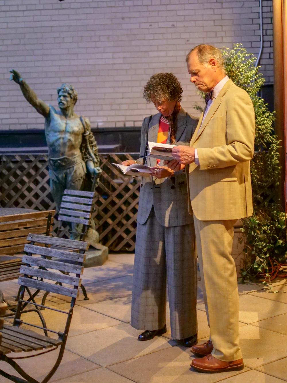 A photo of two people standing to the right of a statue, which is pointing off to the left of the photo. Rima, center, wears a grey suit. Paul, right, wears a tan suit. Both Rima and Paul are holding brochures and reading them.