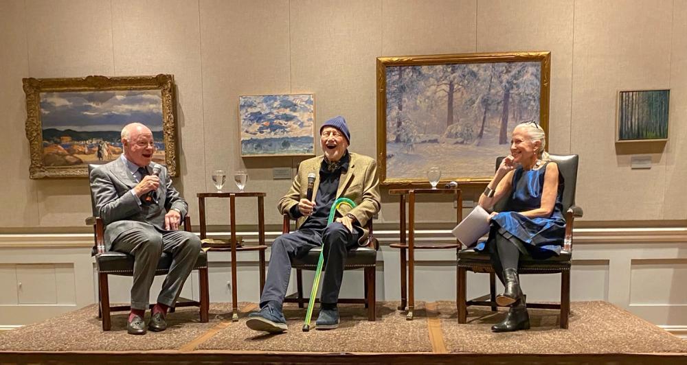 A photo of three people sitting in chairs on a small stage. On the left, Nick is wearing a grey suit and holding a microphone. In the center, Frederic is wearing a navy beanie and a brown blazer with blue pants. He is holding a microphone and a green cane. Erika, on the right, is wearing a blue dress and holding a set of papers. All three are smiling.