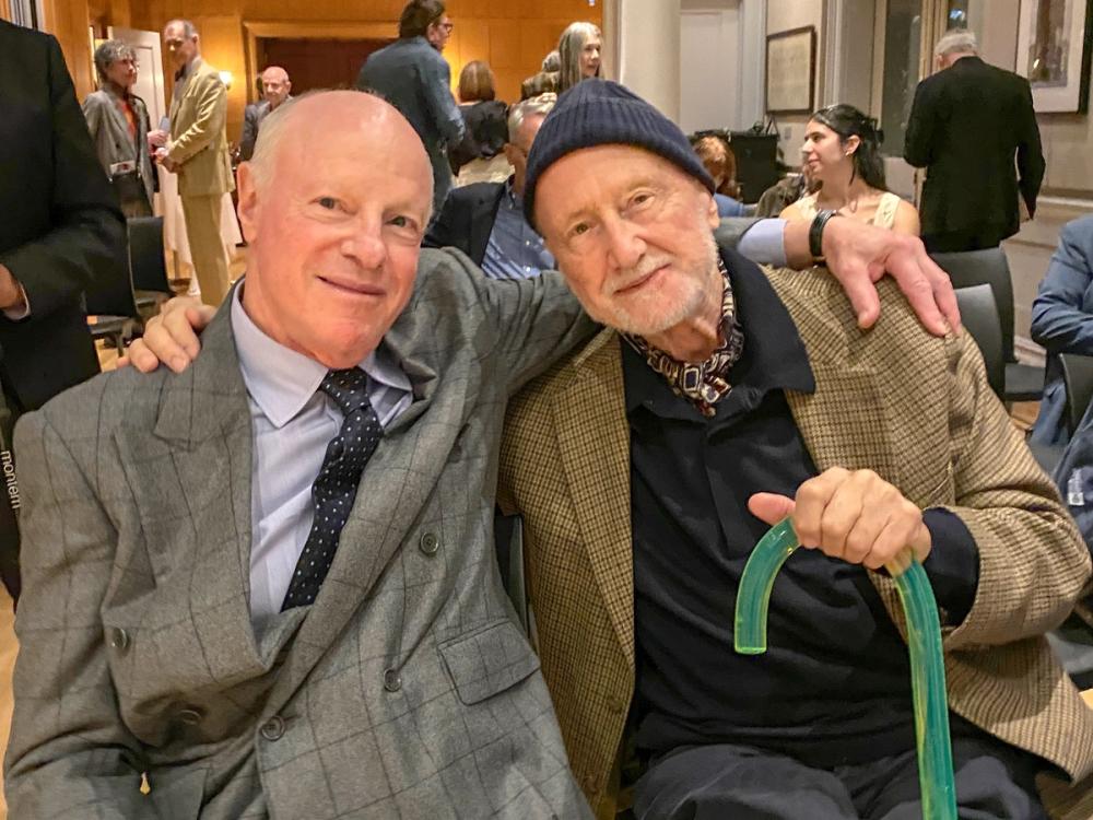 A photo of two men seated with their arms around each other's shoulders. On the left, Nick wears a grey patterned suit jacket and a dark blue polka dot tie. On the right, Frederic is wearing a navy blue beanie and a brown plaid blazer, and he is holding a green cane.