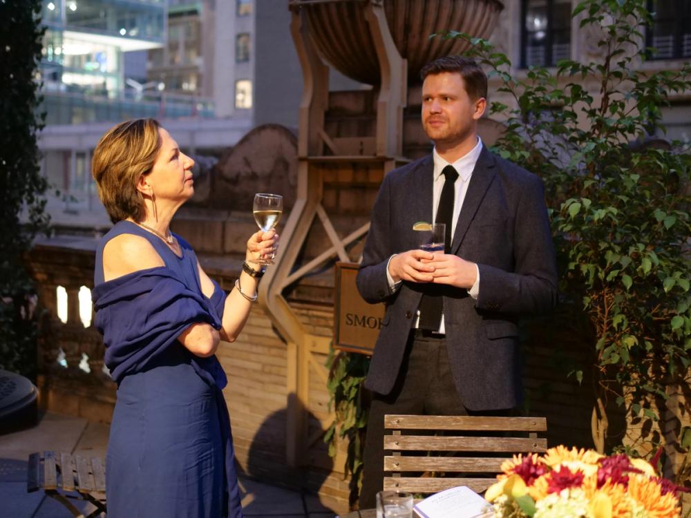 A photo of two people standing on an outdoor terrace. Emma, left, wears a dark blue dress and matching shawl, and she is holding a wine glass in her left hand. Josh, right, is wearing a dark blue blazer and is holding a glass with both hands.