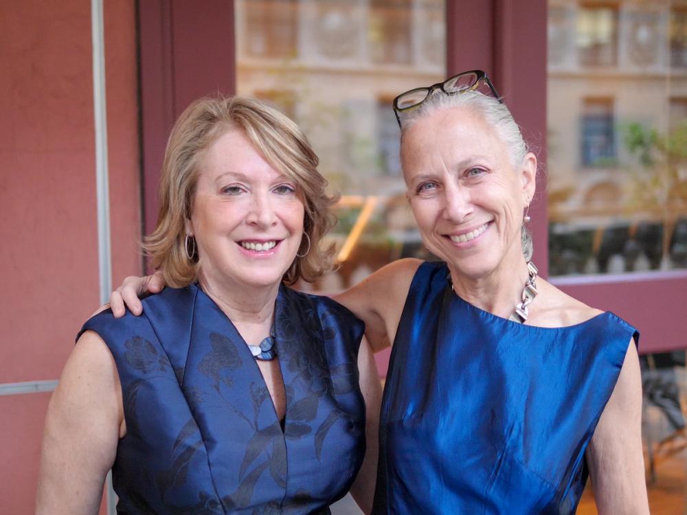 A photo of two women smiling at the camera. Pamela, on the left, wears a dark blue and black dress. Erika, on the left, wears a bright blue dress and has her right arm around Pamela's shoulder.