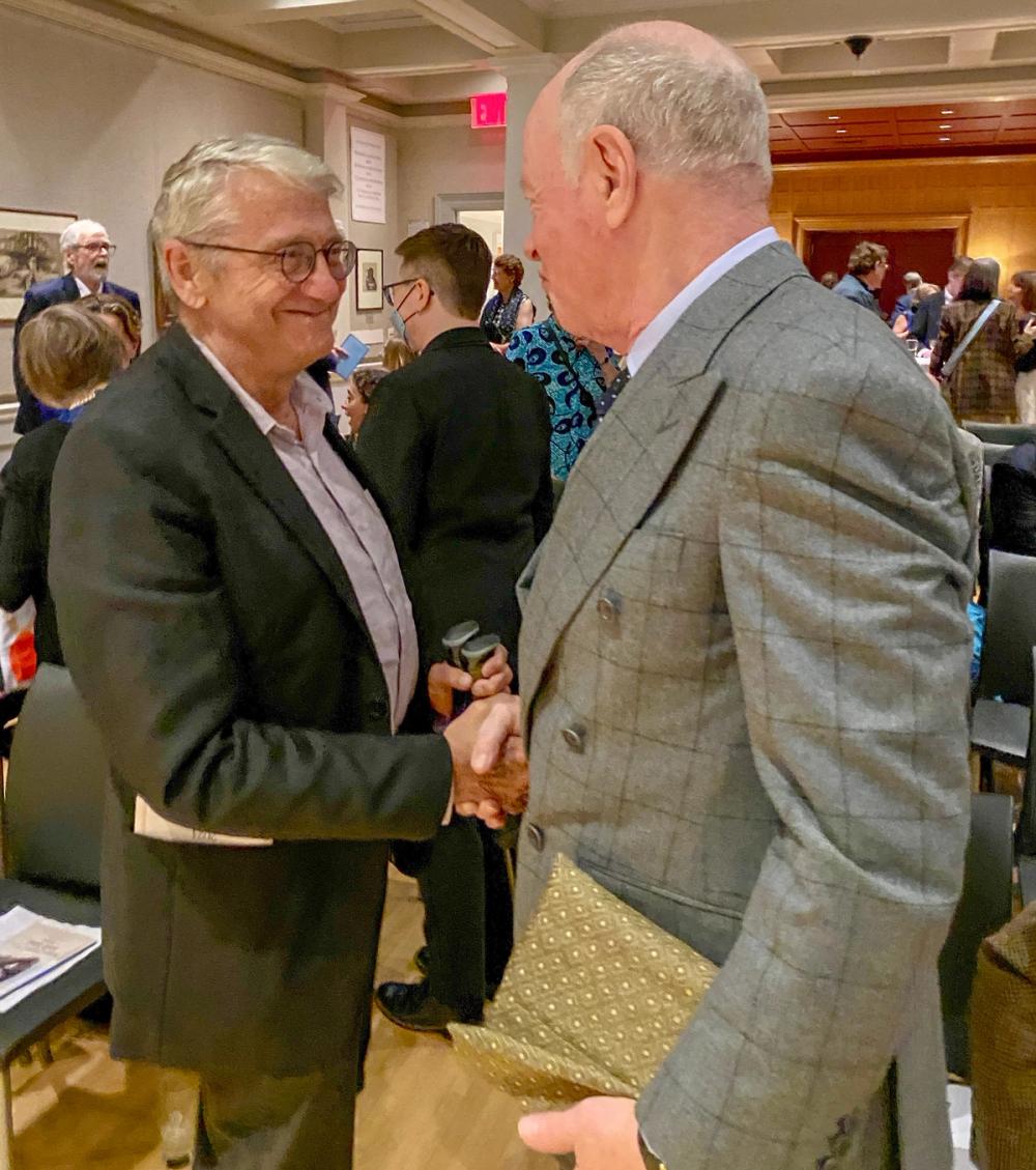 A photo of two men shaking hands and smiling. Steven, left, wears a black blazer and glasses. Nick, right, wears a grey suit jacket and holds a package wrapped in gold paper in his left hand.