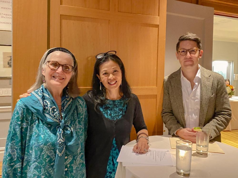 A photo of three people smiling at the camera. Kathleen, left, wears a turquoise blouse and turquoise scarf. Andrea, center, wears a blue and black dress with a black cardigan. Her left arm rests on a high top table. Charlie, right, is wearing a tan blazer and is resting both hands on the high top table.