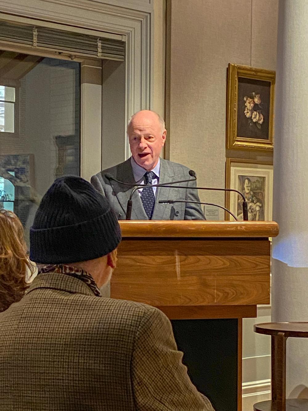 A photo of a man standing at a podium. Nick is wearing a grey suit jacket and a blue polka dot tie.