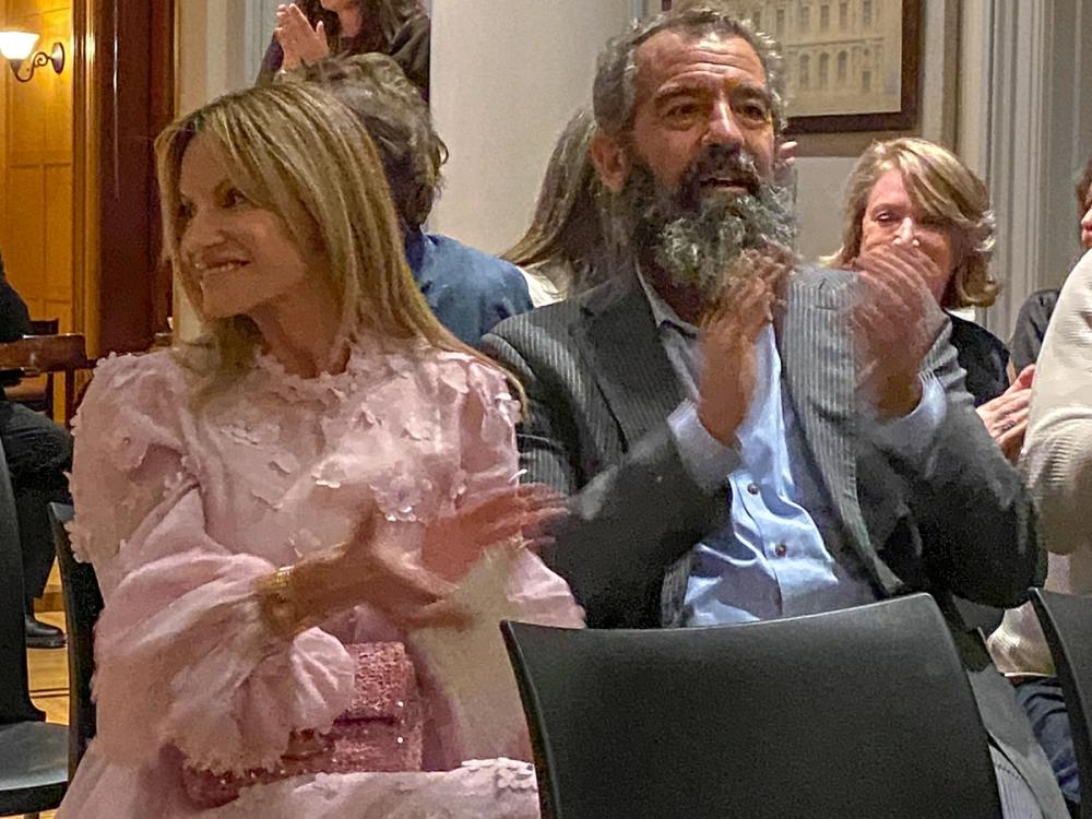 A photo of two people seated and applauding. Lucy, on the left, wears a sparkly light pink dress. Charles, on the right, wears a grey striped blazer and blue button-down shirt.