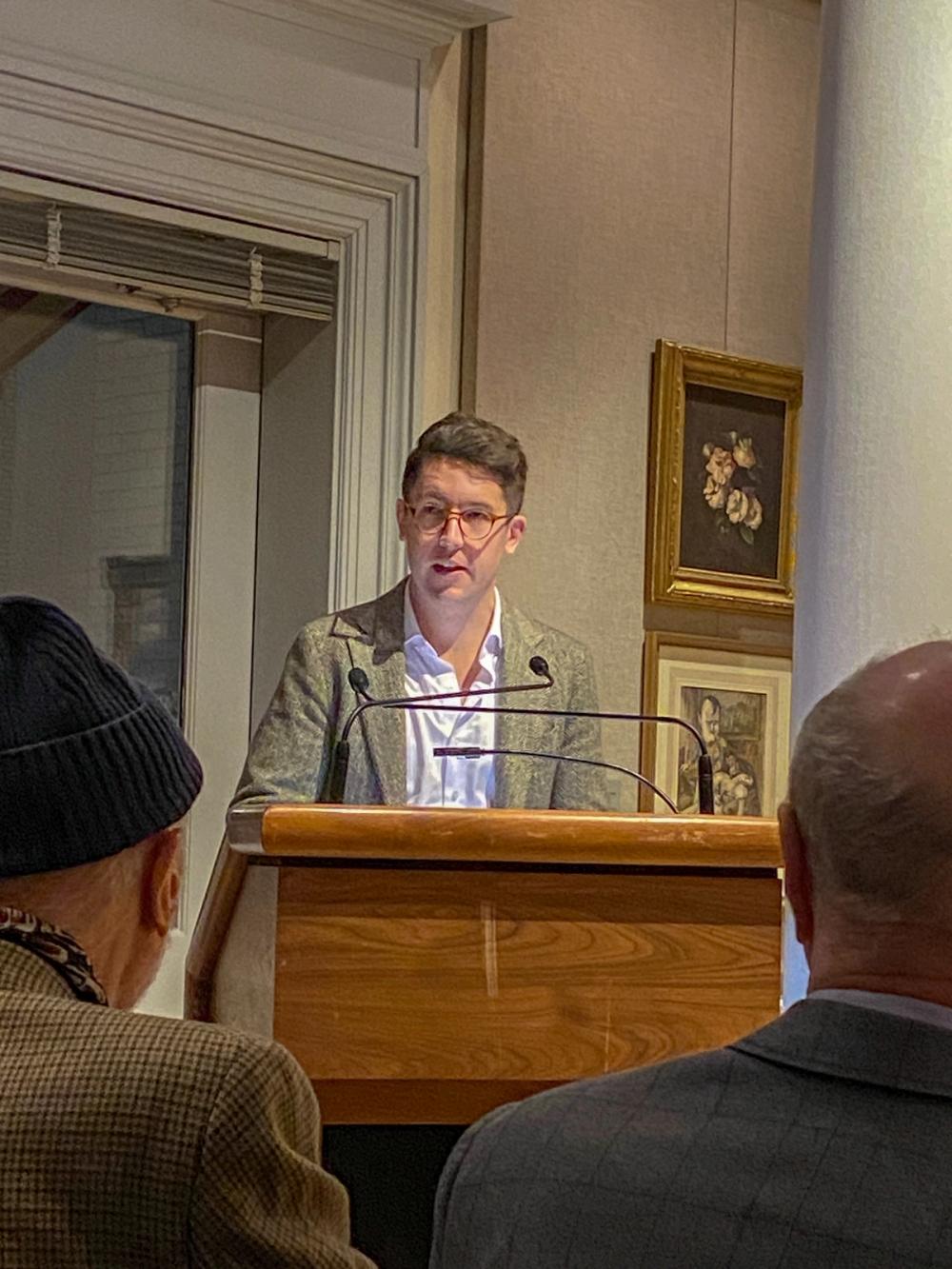 A photo of a man standing at a podium. He wears glasses and a tan blazer.