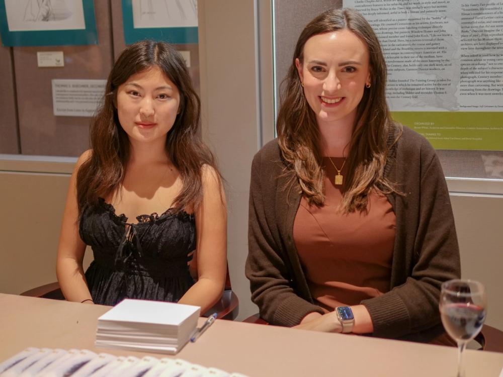 A photo of two women smiling while seated at a table. Audrey, left, wears a black lace dress. Laura, right, wears a brown dress and brown cardigan.