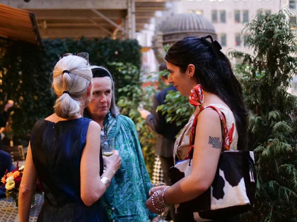 A photo of three women in conversation. Erika, left, has her back turned toward the camera and is wearing a blue dress. Kathleen, center, is wearing a turquoise blouse. Mabel, right, is wearing a white blouse with a cow print bag on her left shoulder.