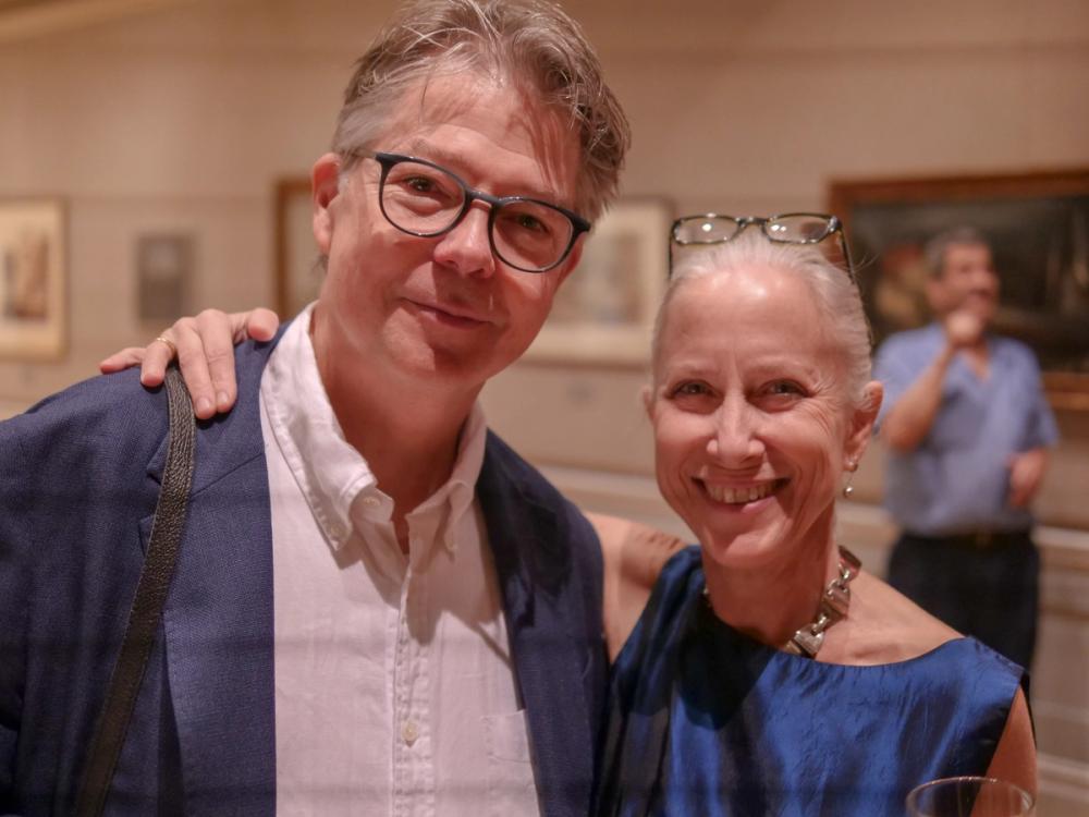 A photo of two people smiling at the camera. Bud, left, wears a blue blazer and white button-down shirt. Erika, right, wears a blue dress and has her right arm over Bud's shoulder.