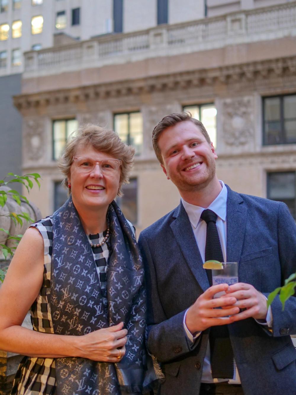 A photo of two people smiling at the camera. Nancy, left, wears a black and white checked dress and a blue Louis Vuitton scarf. Josh, right, wears a blue blazer and a black tie, and is holding a glass with both hands.