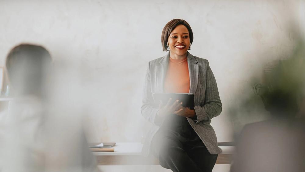 A smiling woman in a grey blazer holding a tablet, seated on the edge of a desk