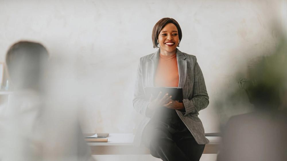 A smiling woman in a grey blazer holding a tablet, seated on the edge of a desk