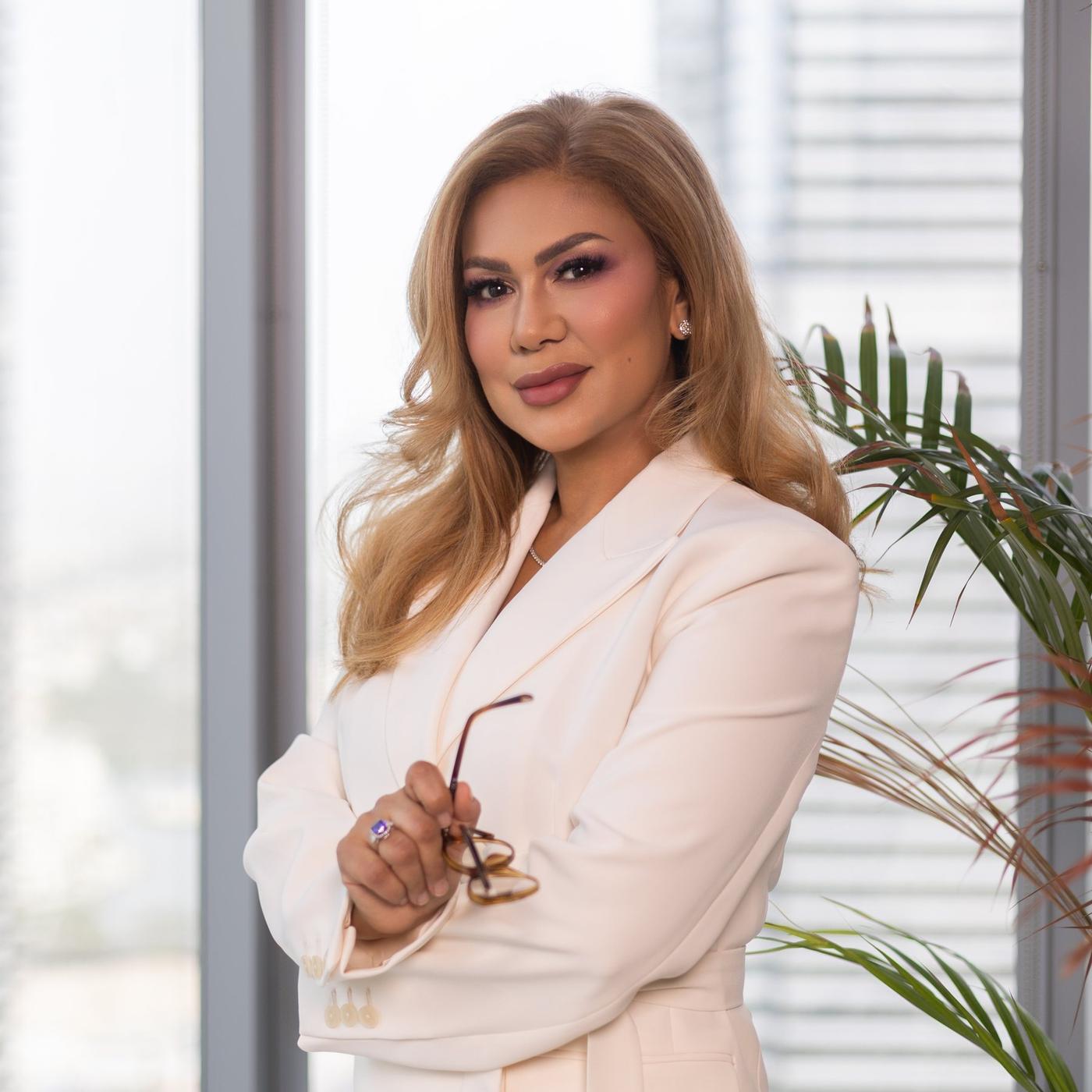 Wardah Harharah in a cream blazer beside floor-to-ceiling windows, holding her glasses, the Dubai skyline behind her