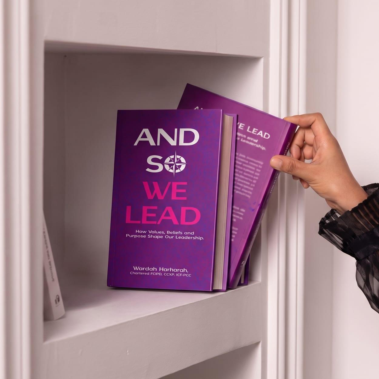 A hand taking a copy of And So We Lead from a white alcove shelf