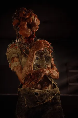 A close up of a clicker standing in a dim room. It seems to be resting, with its arms gently folded around itself. A shaft of light through the blinds makes it easy to make out the tattered clothes and fungal growths all over it.